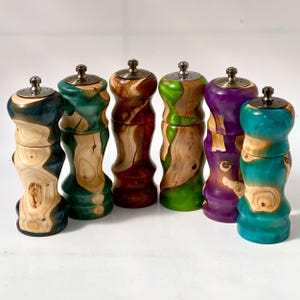 May include: Six cylindrical salt and pepper mills arranged in a row, each featuring a unique combination of natural light-colored wood grain and vibrant resin in shades of blue, green, red, and purple, topped with a metallic grinding mechanism.