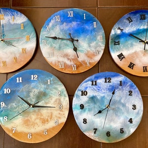 Puede incluir: Cuatro relojes redondos de resina con diseños de olas oceánicas. Cada reloj tiene marcas de números romanos y un esquema de color diferente de azul, blanco y marrón.