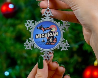 May include: A silver snowflake ornament with a blue and white image of the state of Michigan and the word "MICHIGAN" on it. The ornament is being held by a hand with green nail polish.
