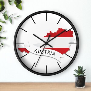 May include: A black wall clock with a white face and black hands. The clock face features a white outline of Austria with the Austrian flag colours, red and white, inside the outline. The word "Austria" is written in black on a white banner.