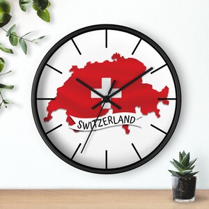 May include: A black framed wall clock with a white face and black hour and minute hands. The clock face features a red and white Swiss flag with the outline of Switzerland in the shape of the flag. The text "SWITZERLAND" is written in a white banner below the flag.