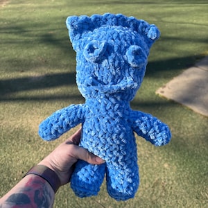 May include: A bright blue plush toy made of soft, chunky yarn. The figure features a rectangular head with a textured top edge, two circular eyes, and a simple stitched mouth. It has a compact body with two arms extending to the sides and two legs.
