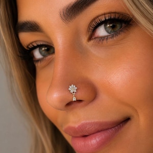May include: A close-up of a nose with a gold-coloured floral nose ring. The nose ring has a flower design with small, clear gemstones and a dangling gemstone. The skin tone is light with a slight tan. The image is well-lit.