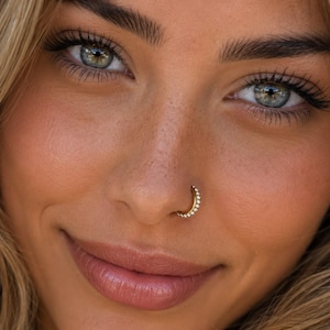Peut inclure: Un anneau de nez doré orné d'une rangée de petites pierres rondes et transparentes incrustées sur le bord extérieur. Le bijou est de forme circulaire et conçu pour être porté comme un piercing dans la narine, avec une finition métallique polie.