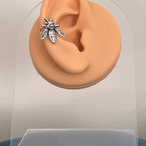 Puede incluir: Pendiente de oreja de abeja de plata. La abeja está detallada con alas, patas y un cuerpo segmentado. El pendiente de oreja se muestra en una oreja modelo.