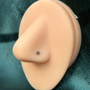 May include: A close-up of a nose piercing on a mannequin. The piercing is a small silver star.