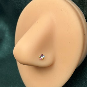 May include: A close-up of a nose piercing with a small flower-shaped stud. The flower has five petals and is adorned with colorful gemstones.