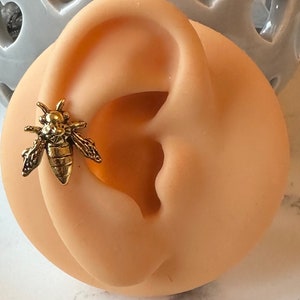 Pode incluir: Piercing de cartilage da orelha em forma de abelha dourada. A abelha é detalhada com asas, pernas e um corpo.