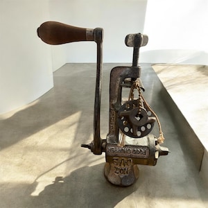 May include: A vintage cast iron food chopper with a wooden handle. The antique kitchen tool features a clamp for securing to a surface, a turning handle, and a grinding mechanism. The metal has a weathered, aged appearance.