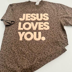 May include: Brown t-shirt featuring a leopard print design. The words "JESUS LOVES YOU" are printed in large, light-coloured letters across the front. The shirt has short sleeves and a crew neck.