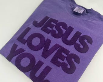 May include: A purple t-shirt with the text "JESUS LOVES YOU" in a dark purple color.