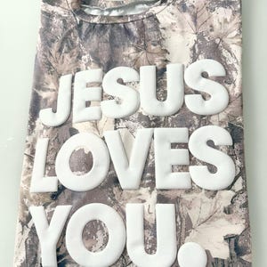 May include: A camouflage-patterned t-shirt with the words "JESUS LOVES YOU" in large, white, puffy letters. The shirt has a crew neck and a neutral color palette of browns, tans, and grays, with a leaf pattern.
