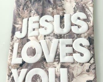 May include: A camouflage-patterned t-shirt with the words "JESUS LOVES YOU" in large, white, puffy letters. The shirt has a crew neck and a neutral color palette of browns, tans, and grays, with a leaf pattern.