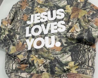 Puede incluir: Una sudadera con estampado de camuflaje con el texto "JESUS LOVES YOU" en letras blancas grandes. La sudadera tiene cuello redondo y mangas largas. El diseño de camuflaje incorpora tonos de marrón, verde y beige, con patrones de hojas y troncos de árboles.