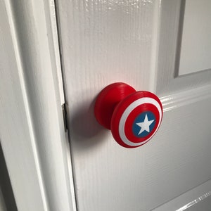 May include: A red and white Captain America shield-themed doorknob is attached to a white door. The doorknob has a red base and a shield design with a white circle, red border, blue accents, and a white star.