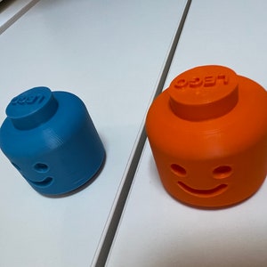 May include: Two 3D printed LEGO minifigure heads, one blue and one orange, with a smiling face and the word "LEGO" on top.