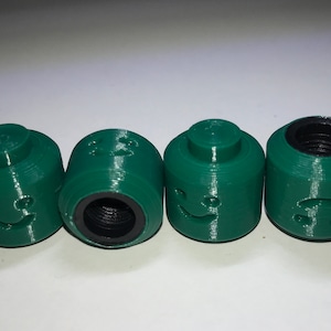 May include: Four green plastic bottle caps with different facial expressions. Each cap has a threaded opening on the bottom.