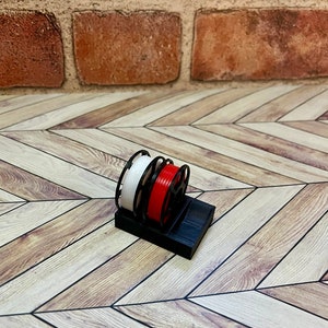 Puede incluir: Un soporte de carrete de pl&aacute;stico negro con dos carretes de filamento, uno blanco y uno rojo.