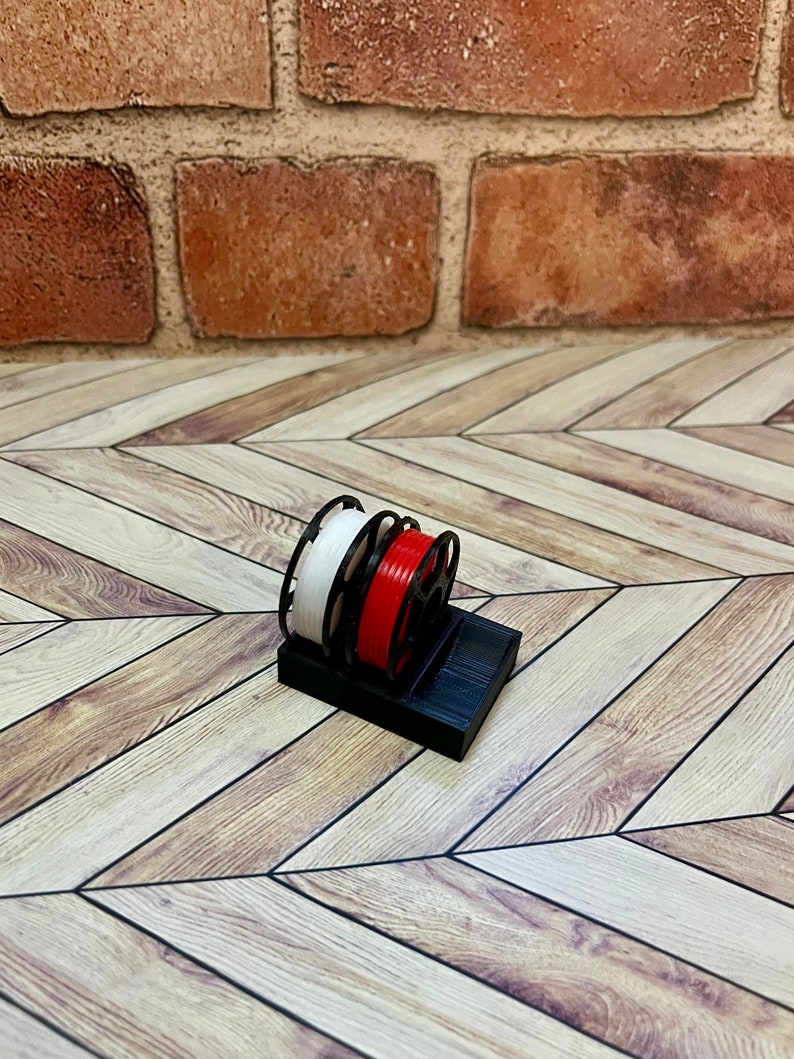 Puede incluir: Un soporte de carrete de pl&aacute;stico negro con dos carretes de filamento, uno blanco y uno rojo.