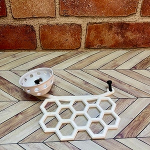 Puede incluir: Plato blanco de pl&aacute;stico en forma de panal con un peque&ntilde;o cuenco blanco de pl&aacute;stico con agujeros. El plato y el cuenco est&aacute;n sobre un suelo de madera.