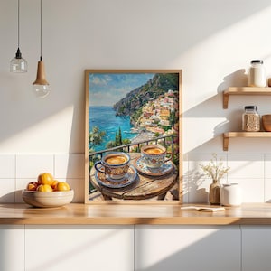 Puede incluir: Un cuadro decorativo enmarcado que muestra dos tazas de caf&eacute; de cer&aacute;mica con motivos sobre una mesa de madera con vistas a una ciudad costera con edificios coloridos, un mar azul y monta&ntilde;as bajo un cielo brillante, en una cocina con armarios blancos.
