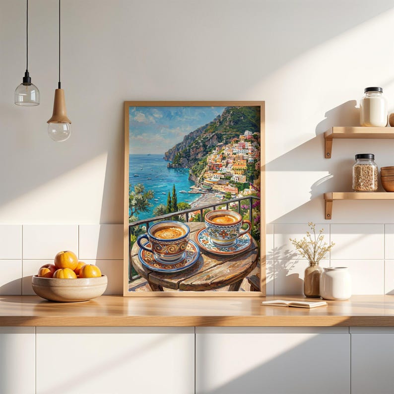 Puede incluir: Un cuadro decorativo enmarcado que muestra dos tazas de caf&eacute; de cer&aacute;mica con motivos sobre una mesa de madera con vistas a una ciudad costera con edificios coloridos, un mar azul y monta&ntilde;as bajo un cielo brillante, en una cocina con armarios blancos.
