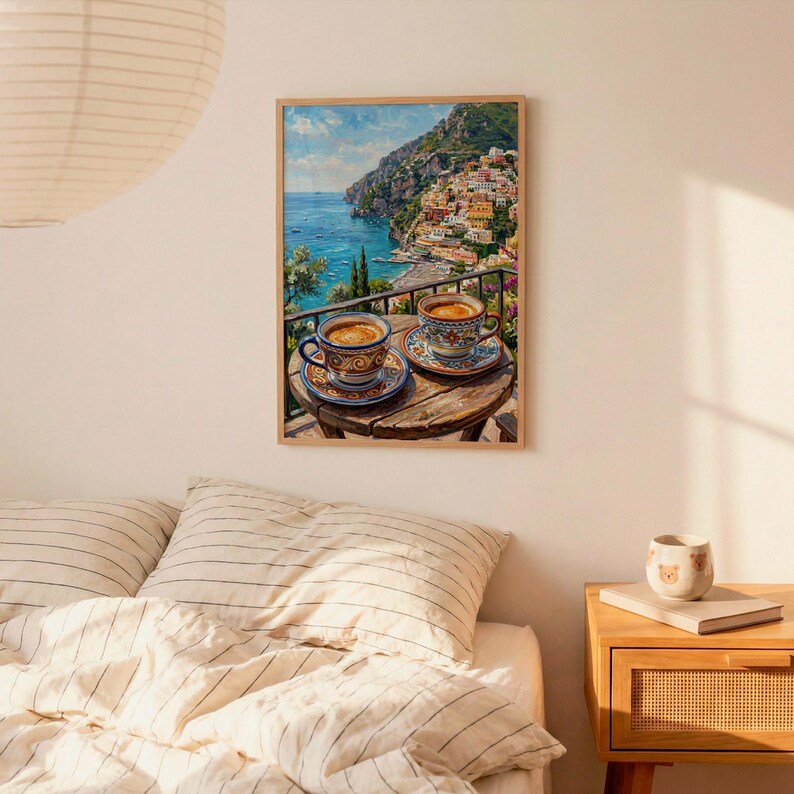 Puede incluir: Una impresi&oacute;n art&iacute;stica enmarcada que representa un paisaje costero con una mesa de madera en primer plano que sostiene dos tazas de caf&eacute; de cer&aacute;mica. La escena muestra un mar azul, un acantilado rocoso y edificios coloridos bajo un cielo soleado.