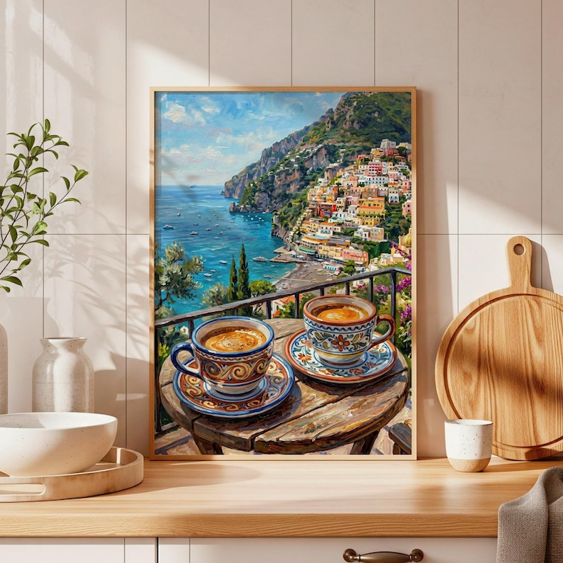 Puede incluir: Una l&aacute;mina de arte enmarcada que representa una escena costera con dos tazas de caf&eacute; de cer&aacute;mica estampadas sobre una mesa de madera con vistas a una ciudad mediterr&aacute;nea en la ladera, el mar azul y el cielo en tonos naranja, azul y verde.