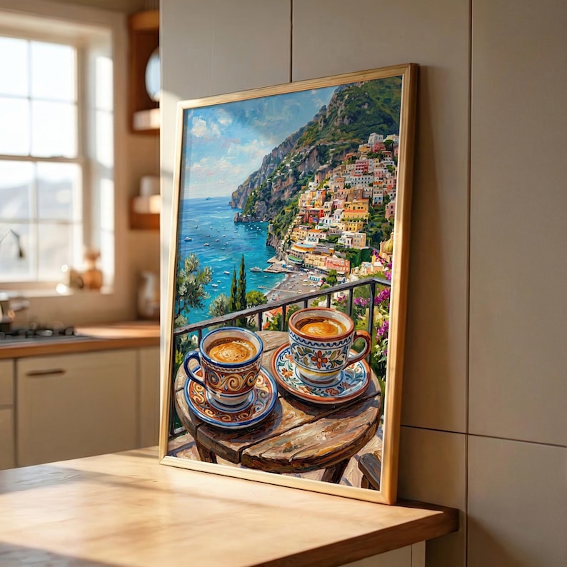 Puede incluir: Una impresi&oacute;n art&iacute;stica enmarcada que representa un pueblo costero italiano en un acantilado sobre el mar, con dos tazas de caf&eacute; de cer&aacute;mica estampadas sobre una peque&ntilde;a mesa redonda de madera en primer plano, frente a una barandilla.