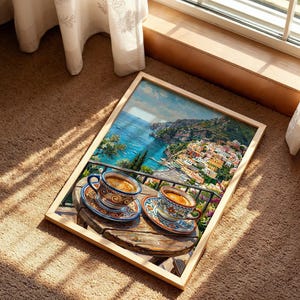 Puede incluir: Una impresi&oacute;n rectangular enmarcada que presenta una vibrante escena costera con dos tazas de caf&eacute; de cer&aacute;mica sobre una mesa de madera. La obra muestra un pueblo mediterr&aacute;neo en la ladera con vistas al mar azul y motivos florales detallados.