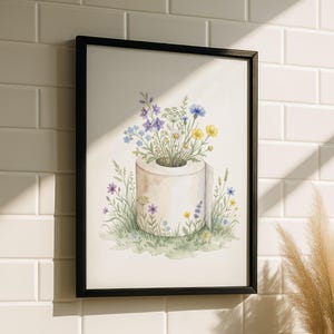 Puede incluir: Una impresión en acuarela enmarcada que presenta un rollo de papel higiénico con un ramo de flores silvestres coloridas. La obra de arte incluye flores azules, amarillas y moradas, sobre un fondo de hierba verde y pared de azulejos blancos.