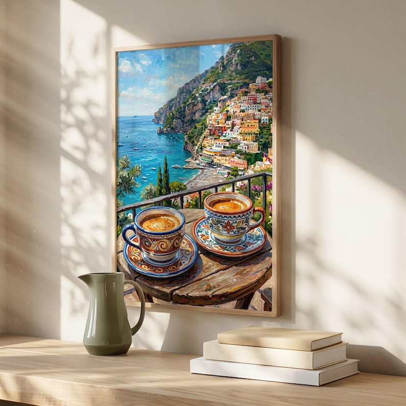 Puede incluir: Una l&aacute;mina de arte enmarcada que representa un pueblo costero italiano en un acantilado sobre el mar, con dos tazas de caf&eacute; de cer&aacute;mica estampadas sobre una mesa de madera, junto a una jarra de cer&aacute;mica verde claro y libros apilados.