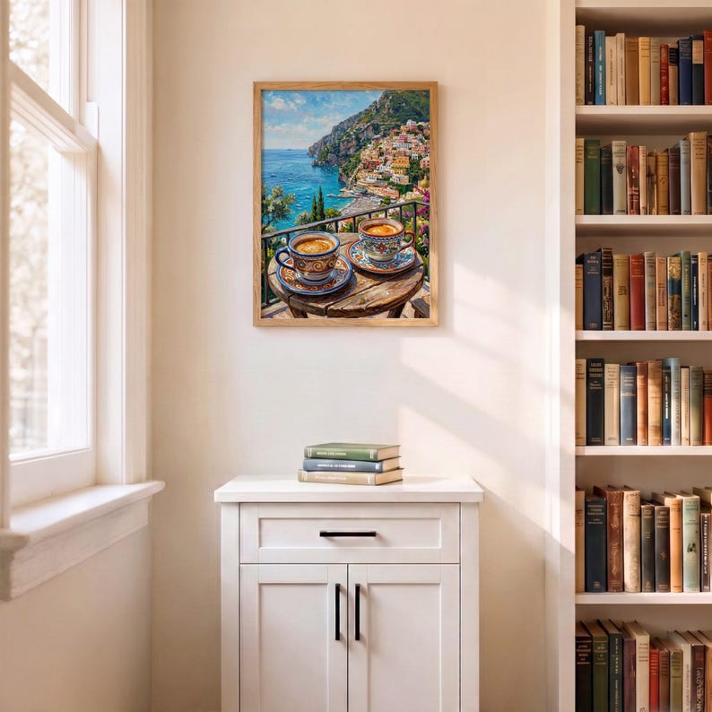 Puede incluir: Una l&aacute;mina de arte enmarcada que representa una escena costera mediterr&aacute;nea con dos tazas de caf&eacute; de cer&aacute;mica estampadas sobre una mesa de madera con vistas a un pueblo en un acantilado, el mar azul y el cielo, sobre un mueble blanco.