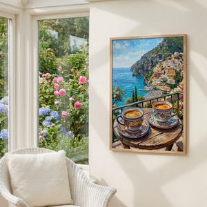 Puede incluir: Una impresi&oacute;n art&iacute;stica enmarcada que representa una escena costera mediterr&aacute;nea con dos tazas de caf&eacute; de cer&aacute;mica estampadas sobre una peque&ntilde;a mesa de madera, con vistas a una ciudad en la ladera con edificios coloridos y un mar azul.