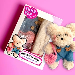 Könnte beinhalten: Ein Bastelset zum Herstellen eines Plüsch-Dungaree-Bären. Das Set enthält einen cremefarbenen Bären, blaue Jeans-Latzhosen, eine rosa gehäkelte Tasche und eine Brille. Das Set ist in einer weißen Box mit rosa und violetten Akzenten verpackt und trägt die Aufschrift "Polly Pop Crafts" und "Dungaree Bear Moru Doll Craft Kit".