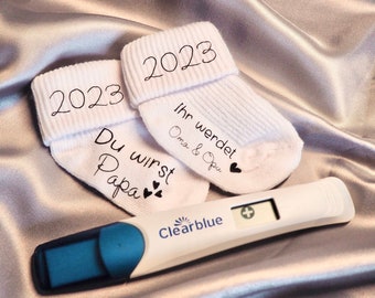 May include: Two white baby socks with the year 2023 printed on the cuffs. One sock features the text Du wirst Papa with three hearts, and the other features the text Ihr werdet Oma & Opu with a heart. A digital Clearblue pregnancy test shows a plus sign.