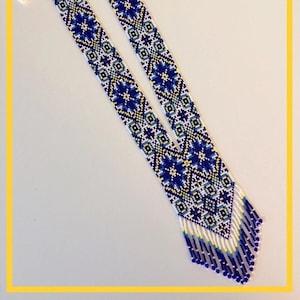Peut inclure: Un long collier de perles blanches avec un motif géométrique bleu, jaune et blanc. Le collier a une frange en bas.