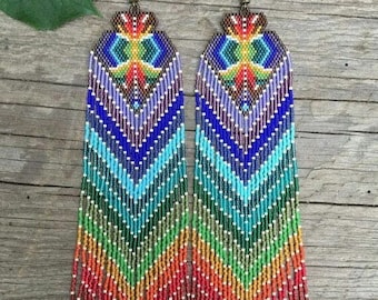 Puede incluir: Un par de aretes largos y coloridos con cuentas y un patrón geométrico. Los aretes están hechos con una variedad de colores, incluyendo rojo, naranja, amarillo, verde, azul y morado. Los aretes están colgando de una superficie de madera.