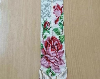 May include: A white beaded necklace with a floral design featuring pink and red roses, green leaves, and gray accents. The necklace has a long fringe at the bottom.