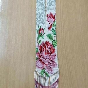 Peut inclure: Un collier de perles blanches avec un motif floral composé de roses roses et rouges, de feuilles vertes et d'accents gris. Le collier a une longue frange en bas.