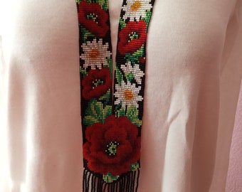 May include: A long beaded necklace with a black background and a floral pattern of red poppies and white daisies. The necklace has a fringe at the bottom.
