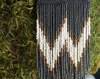 May include: A pair of long, black, white, and gold beaded earrings with a geometric pattern. The earrings are hanging from a branch.