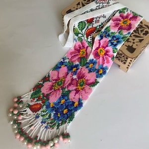 Può includere: Una collana di perline bianche con un motivo floreale in rosa, blu, verde e giallo. La collana ha una frangia di perline bianche.