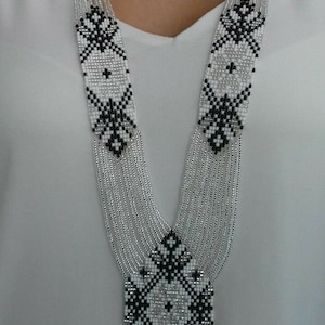 May include: A long, beaded necklace with a geometric pattern in black and white. The necklace has a fringe at the bottom and is made of silver beads.