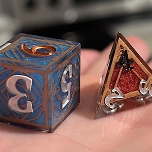 Puede incluir: Dos dados con diseños intrincados. Un dado es un cubo con un diseño azul y cobre y el número 3 en un lado. El otro dado es una pirámide con un diseño rojo y cobre y el número 4 en un lado.