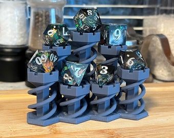 Pode incluir: Um conjunto de seis dados iridescentes com redemoinhos pretos e verdes, exibidos em uma torre azul impressa em 3D com um design em espiral. Os dados são numerados 3, 6, 7, 8, 9 e 0.