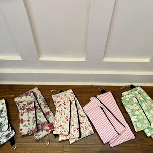 May include: A collection of fabric pouches with various floral and patterned designs. The pouches are in shades of white, pink, green, and red, with black zippers and keychains. The pouches are arranged on a wooden surface.