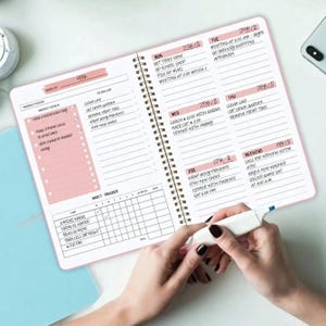 Può includere: Un'agenda a spirale rosa e bianca è aperta su una pagina settimanale. Include sezioni per gli obiettivi settimanali, una lista di cose da fare e un rilevatore di abitudini. Una persona tiene un evidenziatore bianco sopra l'agenda.