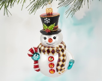 May include: A snowman Christmas ornament with a black top hat, a patterned scarf, and a candy cane. The snowman has a white body with red, orange, and gold buttons. The ornament is hanging from a green pine branch.