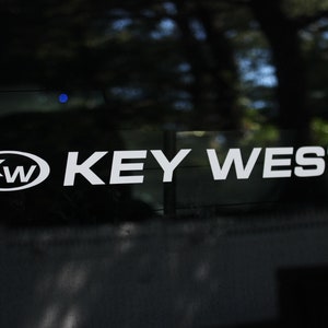 May include: White "KEY WEST" decal on a dark surface. The decal includes the letters "KW" inside an oval. The background is blurred, suggesting an outdoor setting.